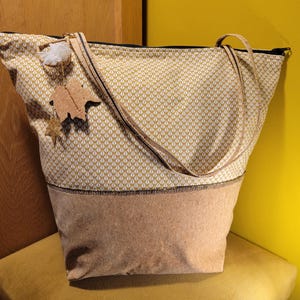 Op de afbeelding: Een tote bag met een geometrisch patroon in crème en goud, en een kurken basis. De tas heeft lange hengsels en een ritssluiting. Een bedel met een blad en een ster hangt aan de rits. De tas staat op een gele stoel.