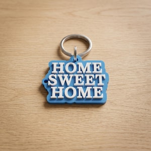 May include: A blue keychain with the words "HOME SWEET HOME" in white lettering. The keychain has a silver ring attached to it. The background is a light brown wooden surface.