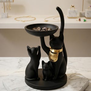 May include: Black cat-shaped jewelry holder with a gold collar, holding a tray with rings. Two smaller black cat figurines are at the base. The holder is on a marble surface, with jewelry displays in the background.