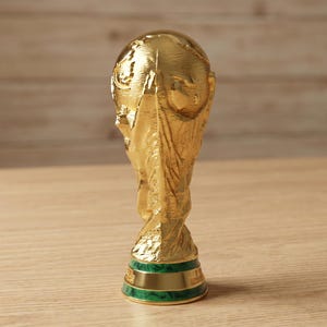 May include: A gold trophy with intricate detailing, including a globe-shaped top and a twisted base. The trophy has a green band at the base and is set against a light wooden backdrop. A symbol of victory and sporting excellence.