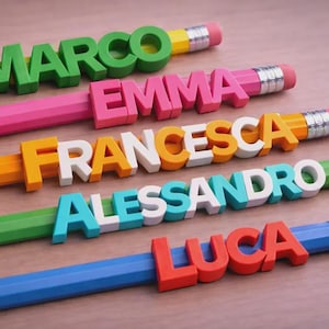 May include: Colorful pencils with names: Marco, Emma, Francesca, Alessandro, and Luca. Each pencil is a different color, with the name spelled out in 3D letters. The pencils have erasers and silver metal bands.