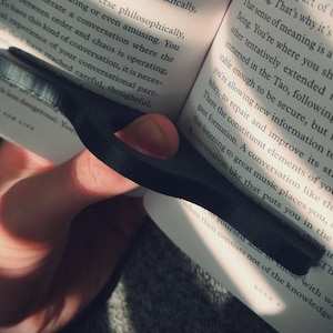 May include: A black, 3D-printed book page holder is shown, designed to keep a book open. The holder is placed on a book with text visible. The design features a curved shape with a finger hole for easy use.