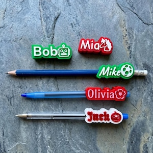 Op de afbeelding: Een verzameling gepersonaliseerde potlood- en penopzetstukken. De opzetstukken zijn in verschillende kleuren en voorzien van namen als "Bob", "Mia", "Mike", "Olivia" en "Jack". Elk opzetstuk heeft een uniek ontwerp, waaronder een robot, een eenhoorn, een voetbal, een bloem en een pompoen.