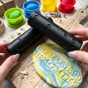 Peut inclure: Deux rouleaux cylindriques noirs présentant des chiffres en relief de zéro à neuf. Un rouleau est pressé dans un disque de pâte marbrée bleue, jaune et blanche, laissant une empreinte numérique claire. L'ensemble inclut des contenants en plastique.