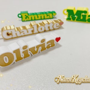 Könnte beinhalten: Vier 3D-gedruckte Namensschlüsselanhänger mit den Namen Olivia, Charlotte, Emma und Mia. Die Schlüsselanhänger sind in den Farbkombinationen Weiß und Gold oder Grün und Gelb gehalten und jeweils mit einem kleinen Herzsymbol versehen.