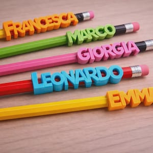 May include: A close-up of five pencils with colorful name tags spelling out "FRANCESCA", "MARCO", "GIORGIA", "LEONARDO", and "EMMA". The pencils are yellow, green, pink, red, and yellow. The name tags are orange, green, pink, blue, and orange.