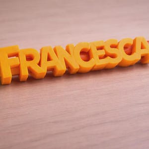 May include: A yellow pencil with the name "FRANCESCA" in orange 3D letters. The pencil has a pink eraser and silver metal band. The name is centered on the pencil, creating a personalized stationery item.