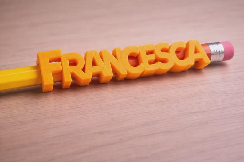 May include: A yellow pencil with the name "FRANCESCA" in orange 3D letters. The pencil has a pink eraser and silver metal band. The name is centered on the pencil, creating a personalized stationery item.