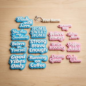 May include: A collection of colorful, word-shaped keychains and charms. The keychains feature phrases like "Made with Love," "You Are My Sunshine," and "Good Vibes Only." The charms are in blue and pink with white lettering.
