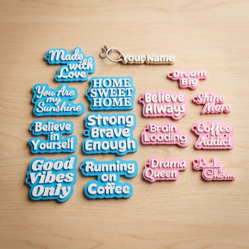 May include: A collection of colorful, word-shaped keychains and charms. The keychains feature phrases like "Made with Love," "You Are My Sunshine," and "Good Vibes Only." The charms are in blue and pink with white lettering.