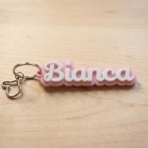 Peut inclure: Un porte-clés rose et blanc avec le nom "Bianca" en écriture cursive. Le porte-clés a un porte-clés argenté et un petit charme en forme de cœur. Le fond est une surface en bois clair.