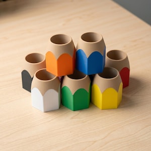 May include: A collection of colorful pencil-shaped desk organizers. Each organizer features a different color, including orange, blue, green, yellow, white, red, and black. The pencil-shaped containers are arranged on a light-colored wooden surface.