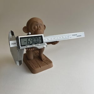 May include: A brown monkey figurine holding a digital caliper. The caliper displays "-6900" on its screen and has a stainless steel ruler with measurements in millimeters. The figurine is on a wooden base.