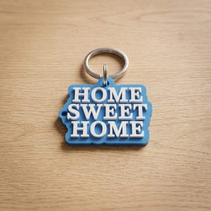 May include: A blue keychain with the words "HOME SWEET HOME" in white, attached to a silver metal ring. The keychain is on a light brown wooden surface.