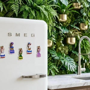 May include: White SMEG refrigerator with colorful magnet decorations. The magnets depict women in traditional dresses. The background features a green wall of plants and gold planters, with a stainless steel faucet and sink.