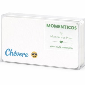 Puede incluir: Una caja de plástico transparente que contiene una pila de tarjetas blancas. Las tarjetas tienen texto en azul y verde, incluyendo la palabra "Chévere" y la frase "Momenticos by Momenticos Press". También se ve un emoji con gafas de sol.