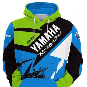 Könnte beinhalten: Ein leuchtend blauer, lindgrüner und schwarzer Kapuzenpullover mit dem Yamaha-Logo und dem Schriftzug "YAMAHA Revs Your Heart" auf der Vorderseite. Das Kleidungsstück verfügt über eine Kängurutasche, weiße Kordelzüge und ein GYTR-Logo.