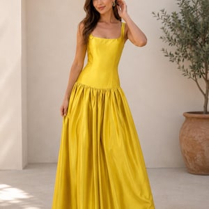 May include: A floor-length, bright yellow satin dress featuring a fitted bodice with a square neckline and thick shoulder straps. The garment has a dropped waistline that transitions into a voluminous, pleated skirt that falls to the floor.
