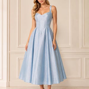 May include: A light blue midi dress with a sweetheart neckline, thick shoulder straps, and a fitted bodice that transitions into a full, pleated skirt. The fabric has a subtle, shimmering metallic finish that catches the light.