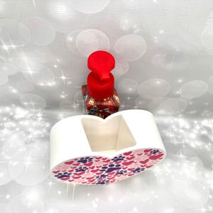 Puede incluir: Un jabonera blanca en forma de corazón con un estampado de huellas y corazones rosas, morados y blancos. Detrás de la jabonera hay un dispensador de jabón con tapa roja. El dispensador tiene un diseño decorativo.