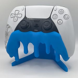 Puede incluir: Un mando blanco y negro de PlayStation 5 con un diseño azul goteante. El mando descansa sobre un soporte azul a juego. El mando tiene la disposición estándar de botones y el logotipo de PlayStation.