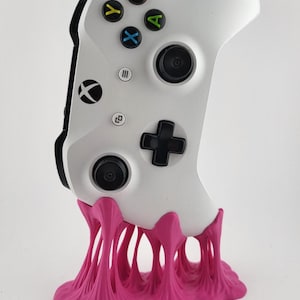 Puede incluir: Un mando blanco de Xbox se muestra sobre un soporte rosa. El mando tiene botones negros y botones de colores A, B, X e Y. El soporte está diseñado para parecer chicle rosa.