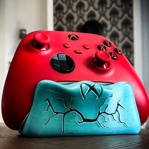 Puede incluir: Un mando Xbox rojo descansa sobre un soporte turquesa con diseño agrietado. El mando tiene botones y joysticks negros. El logotipo de Xbox es visible. Diseño moderno y llamativo, ideal para jugadores.