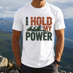 Op de afbeelding: Een wit t-shirt met korte mouwen voorzien van een grafisch ontwerp met de tekst I HOLD MY POWER in blokletters. Het ontwerp bevat een gestileerde illustratie van donkergroene bergtoppen tussen de woorden, met oranje en groene tekst.