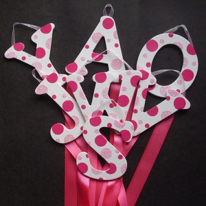 May include: Five white wooden letters with pink polka dots spelling out the word "YOURS" with pink ribbon attached to each letter.