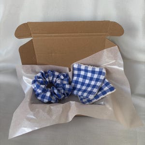Peut inclure: Un chouchou à carreaux bleu et blanc et un carré de tissu assorti sont présentés dans une boîte en carton marron. Les articles sont posés sur du papier de soie blanc, créant une présentation visuellement attrayante.