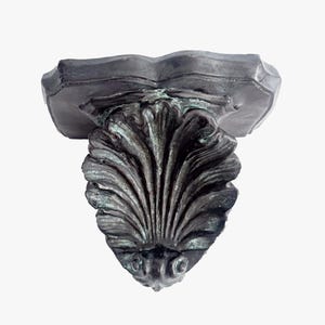 May include: A dark gray, ornate wall shelf with a scalloped top and a large, stylized shell design. The shelf has a weathered appearance with hints of green patina, suggesting an aged, antique aesthetic. The shelf is designed to be mounted on a wall.