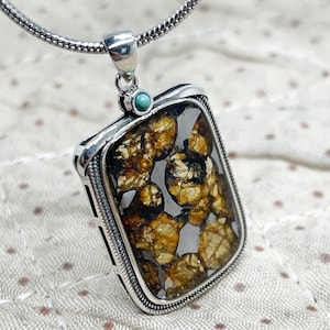 Puede incluir: Un collar con colgante de plata con una piedra rectangular estampada. La piedra presenta tonos marrones y dorados, engastada en un marco plateado. Una pequeña piedra turquesa adorna la parte superior del colgante.