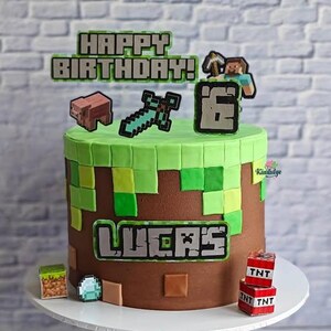 Può includere: Una torta di compleanno rotonda a tema Minecraft. La torta ha una glassa marrone con quadrati verdi e gialli sulla parte superiore. È decorata con personaggi di Minecraft, il numero 6 e il nome "Lucas". Le parole "Happy Birthday!" sono in cima.