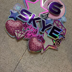 May include: A glittery cake topper with the name "SKYE" in purple, surrounded by stars, hearts, musical notes, and a disco ball design. The number "6" is also included. The topper is on a clear stick.