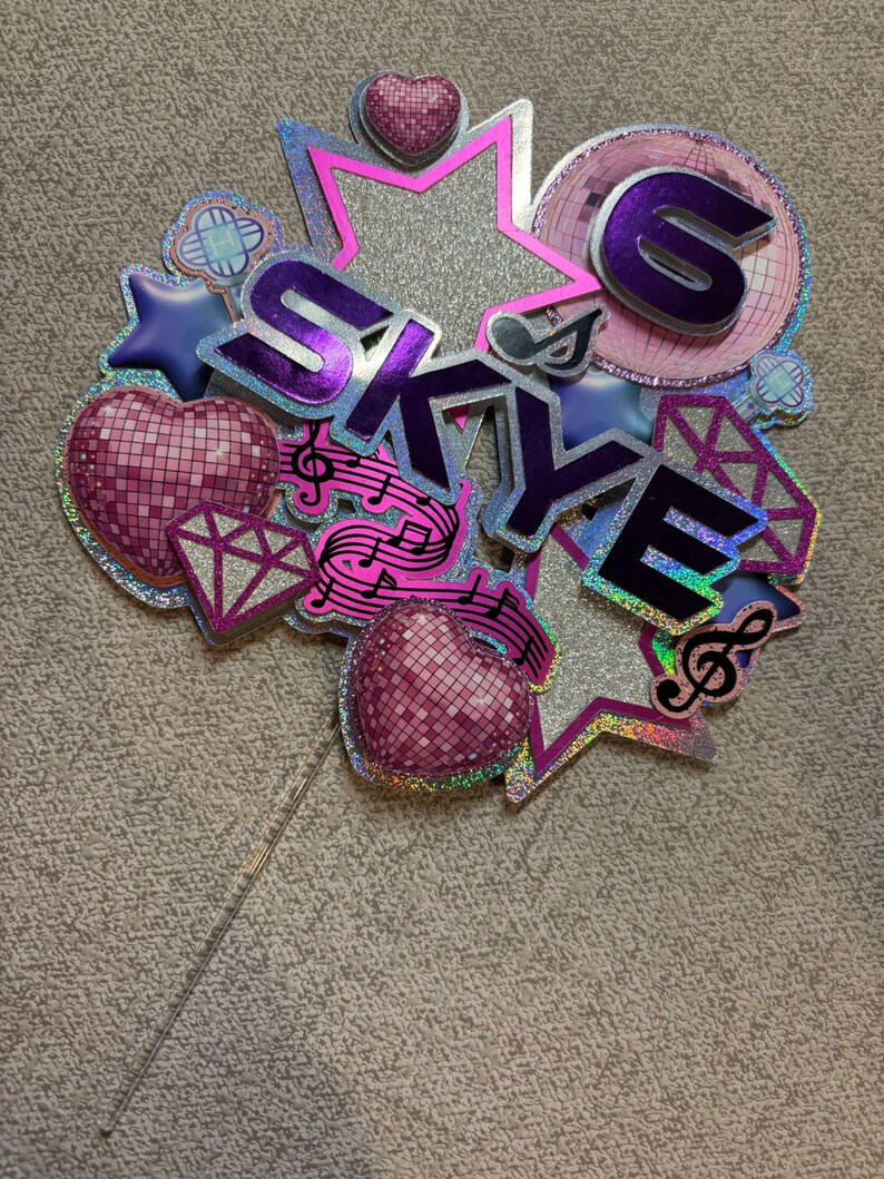 May include: A glittery cake topper with the name "SKYE" in purple, surrounded by stars, hearts, musical notes, and a disco ball design. The number "6" is also included. The topper is on a clear stick.