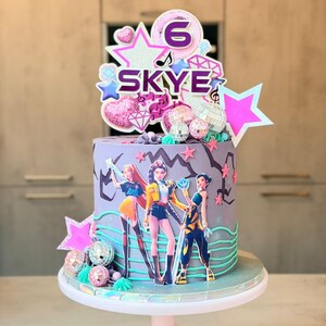 May include: A birthday cake with a purple base, decorated with a pop star theme. The cake features a topper with the name "Skye" and the number "6", along with star and disco ball decorations. Three pop star figures are on the cake.