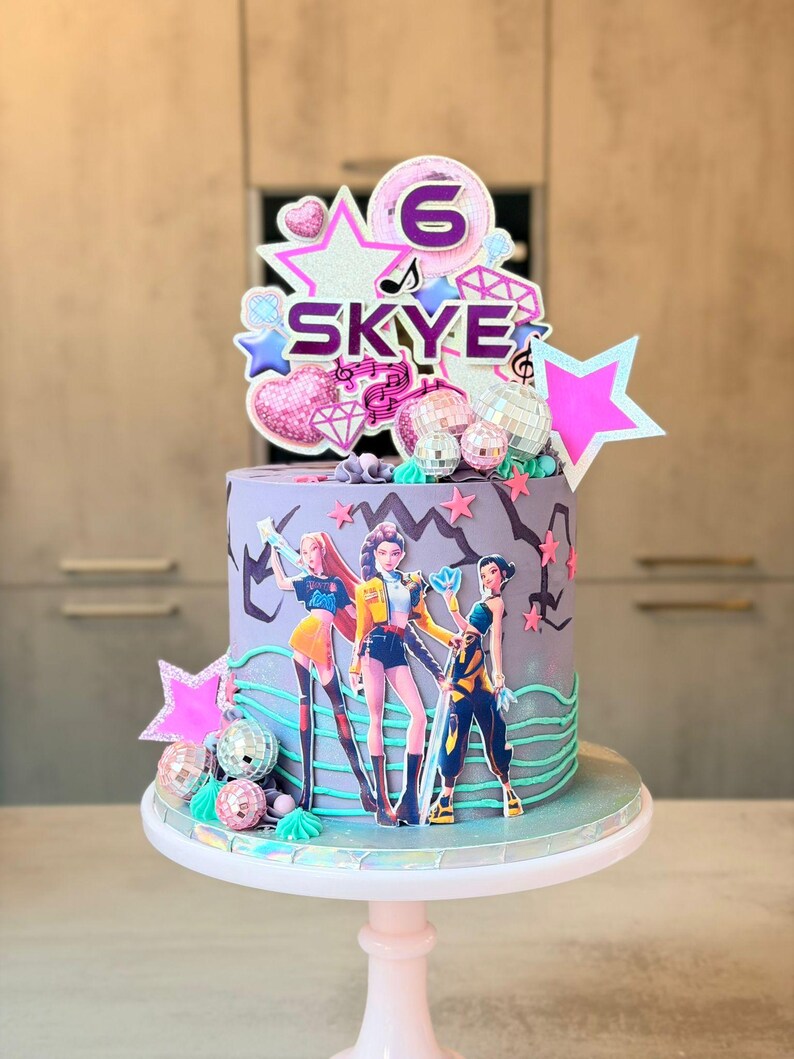 May include: A birthday cake with a purple base, decorated with a pop star theme. The cake features a topper with the name "Skye" and the number "6", along with star and disco ball decorations. Three pop star figures are on the cake.
