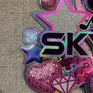 May include: A colorful, glittery decoration with the word "SKY" in purple letters. The design includes a silver star, a pink disco ball heart, a diamond, musical notes, and other decorative elements. The overall theme is celebratory.
