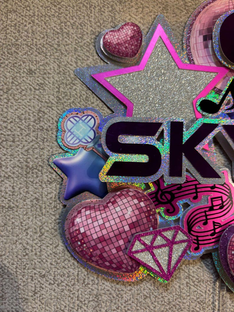 May include: A colorful, glittery decoration with the word "SKY" in purple letters. The design includes a silver star, a pink disco ball heart, a diamond, musical notes, and other decorative elements. The overall theme is celebratory.
