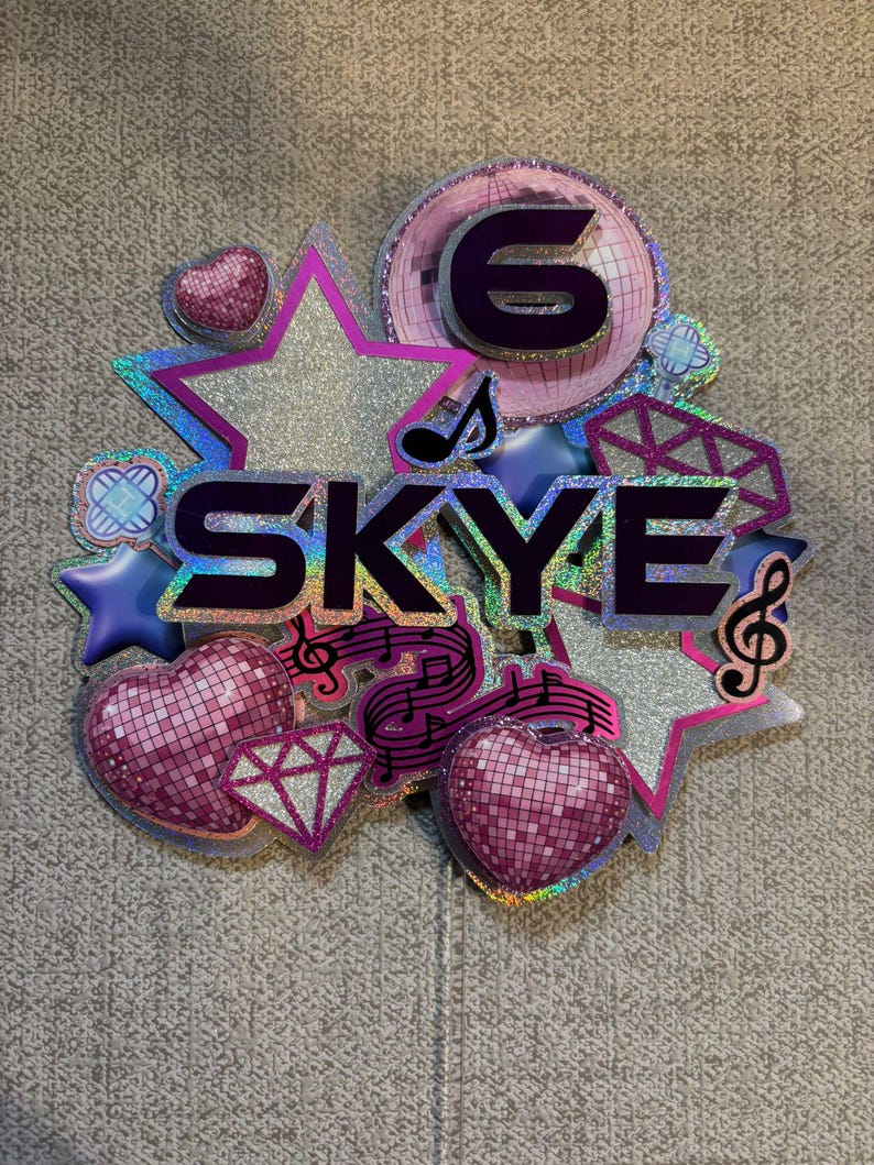 May include: A colorful cake topper with the name "SKYE" and the number "6". The design includes stars, hearts, musical notes, and a disco ball, all in shades of pink, purple, and silver. The topper has a holographic effect.