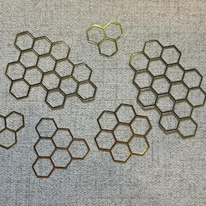 以下が含まれることがあります： さまざまなサイズの金色のハニカム型金属片が、質感のある明るい灰色の表面に配置されています。 各六角形が明確に定義された、オープンフレームの作品です。 この配置は、工芸品または装飾的なテーマを示唆しています。