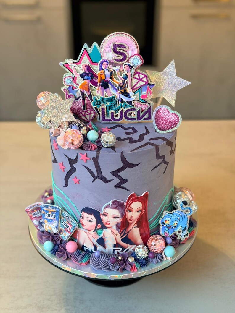 May include: A decorated birthday cake with a purple base and a variety of decorations. The cake features a topper with the number 5 and the name "Luca". Additional decorations include disco balls, stars, and images of cartoon characters.