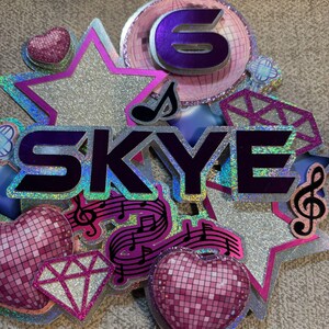 May include: A glittery birthday cake topper with the name "SKYE" in purple letters. The topper features a large number "6", stars, hearts, musical notes, and a diamond shape, all in shades of pink, silver, and purple.