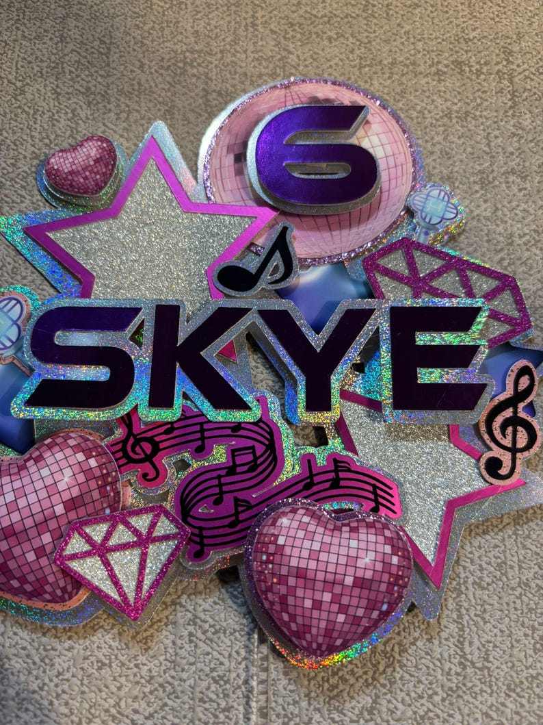 May include: A glittery birthday cake topper with the name "SKYE" in purple letters. The topper features a large number "6", stars, hearts, musical notes, and a diamond shape, all in shades of pink, silver, and purple.