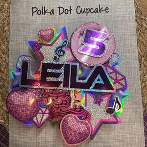 May include: A colorful cake topper with the name "LEILA" in purple letters. The topper includes a number "5", musical notes, stars, and heart-shaped disco balls. The words "Polka Dot Cupcake" are at the top.