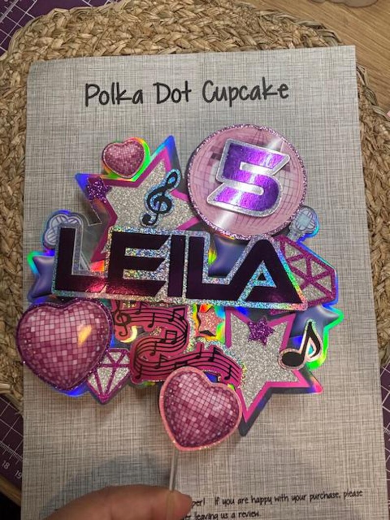 May include: A colorful cake topper with the name "LEILA" in purple letters. The topper includes a number "5", musical notes, stars, and heart-shaped disco balls. The words "Polka Dot Cupcake" are at the top.