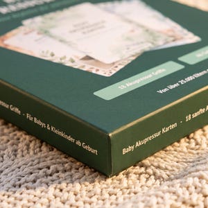 K&ouml;nnte beinhalten: Eine dunkelgr&uuml;ne rechteckige Schachtel mit einem Kartenset. Die Schachtel ist mit wei&szlig;em Text bedruckt, der Baby Akupressur Karten, 18 sanfte Akupressur Griffe und F&uuml;r Babys & Kleinkinder ab Geburt lautet. Die Karten haben einen Blumenrand.