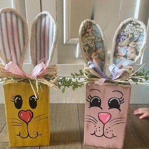 Puede incluir: Dos decoraciones de conejitos de Pascua de madera. Una es amarilla con rasgos faciales rosas y negros y orejas rosas. La otra es rosa con rasgos faciales negros y orejas con estampado floral. Ambas tienen detalles de rafia y cinta.