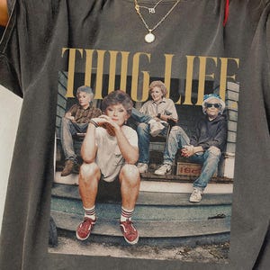 May include: Dark gray t-shirt featuring a graphic of four people sitting on a porch with the words "THUG LIFE" in gold lettering. The graphic is a parody of the Golden Girls. The shirt has short sleeves and a relaxed fit.