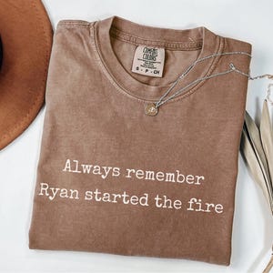 Puede incluir: Una camiseta marrón Comfort Colors con el texto blanco "Always remember Ryan started the fire". Un collar plateado con un colgante está sobre la camiseta. Un sombrero marrón y hojas secas están en el fondo.